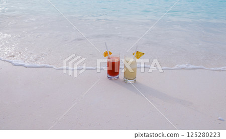 Two colorful delicious cocktails on the ocean. Glasses with cool drinks and pieces of fruit near the clear blue water on the shore. 125280223