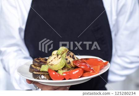 A close-up of a man hand holding a plate of grilled vegetables. A waiter in a white shirt and black apron with a dish of fragrant eggplant, pepper and zucchini. 125280234