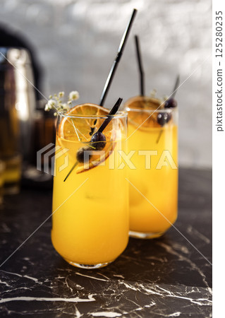 Close-up of two glasses with a citrus cocktail on a black marble table. Tasty chilled drinks are decorated with orange slices, dried flowers and olives. 125280235