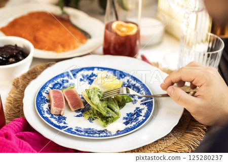 A close-up of a plate with blue decor and pieces of fish and salad. A man hand holds a fork. A tasty fresh tuna dish with lettuce leaves. 125280237