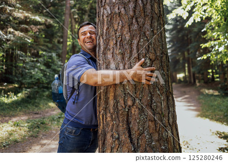 A young man with a backpack in the woods. An attractive tourist touches a large tree in the forest A young man with a backpack in the woods. An attractive tourist touches a large tree in the forest 125280246