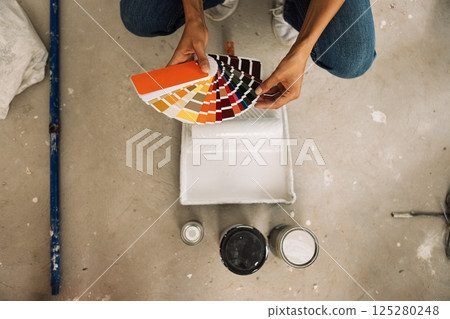 Close-up of female hands holding palette of colors, cans and paint tray on concrete floor. Young woman selects color of paint using paint swatches. Top view. Close-up of female hands holding palette of colors, cans and paint tray on concrete floor. Young woman selects color of paint using paint swatches. Top view. 125280248
