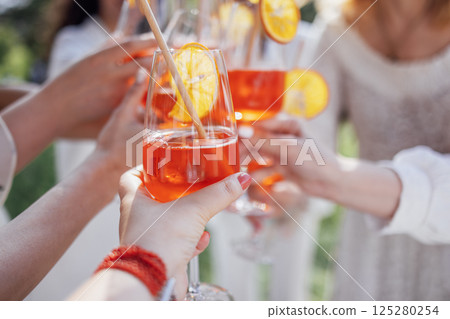 Close-up of a female hands with glasses with a delicious aperol. Young women in white clothes make toast with orange alcoholic drinks. 125280254