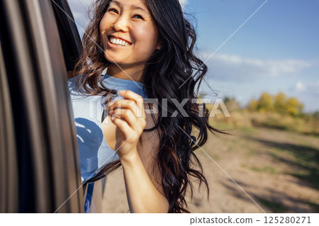 A young Asian look out of the car window and laughs. A smiling Korean woman travels and has fun on the road. A charming girl enjoys the ride and nature. 125280271