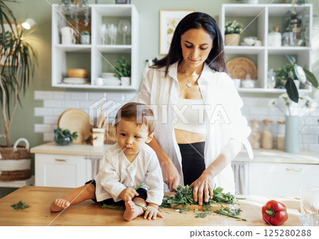 A young mother is cutting greens on a wooden stand in a modern kitchen and her little baby is playing next to her on the table. 125280288
