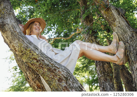 A cheerful young woman in a light summer sundress and a straw hat is laughing sitting on a tree. An attractive blonde is fooling around and having fun in nature. Copy space. 125280306