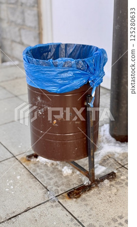A brown dumpster with a blue trash bag is installed on the sidewalk next to the store. Close-up. 125280633