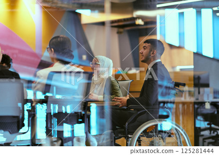 In a modern glass startup office, a wheelchair-bound director leads a successful meeting with colleagues, embodying inclusivity and teamwork. 125281444