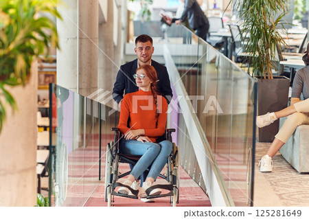 A company director assists his business colleague in a wheelchair, helping her navigate to their startup office, where they work alongside their diverse team of colleagues, emphasizing inclusivity and 125281649