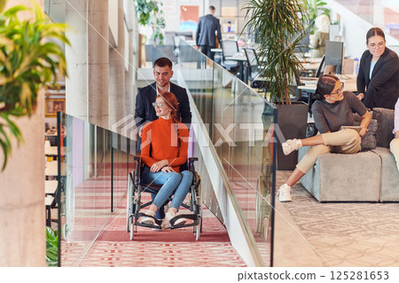 A company director assists his business colleague in a wheelchair, helping her navigate to their startup office, where they work alongside their diverse team of colleagues, emphasizing inclusivity and 125281653