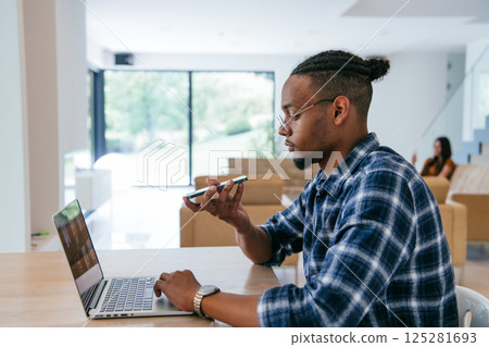 African American businessman in glasses sitting at a table in a modern living room, using a laptop and smartphone for business video chat, conversation with friends and entertainment. African American businessman in glasses sitting at a table in a modern living room, using a laptop and smartphone for business video chat, conversation with friends and entertainment. 125281693