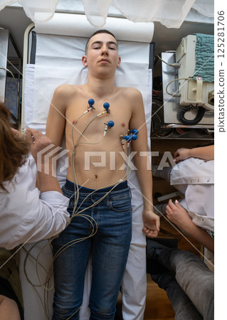 A female doctor performs an ECG on a young man in a hospital room, carefully monitoring his heart health during a routine checkup. 125281706