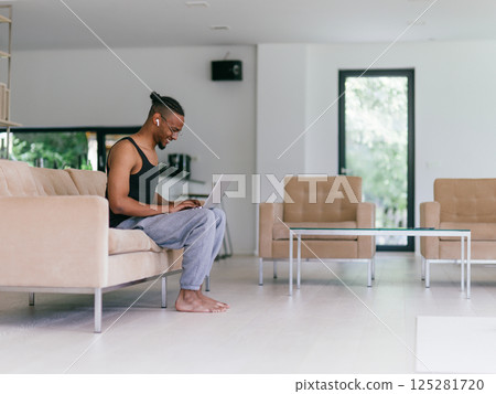 African American man in glasses sitting at a table in a modern living room, using a laptop for business video chat, conversation with friends and entertainment. 125281720