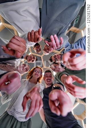 A group of cheerful young people gathered in a circle, pointing at the camera from a low-angle perspective. 125281762