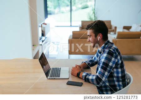 A freelancer sitting at a table in a modern living room, with headphones using a laptop for business video chat, conversation with friends and entertainment A freelancer sitting at a table in a modern living room, with headphones using a laptop for business video chat, conversation with friends and entertainment 125281773
