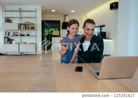 Mother with her daughter talking on laptop with family and friends while sitting in modern living room of big house. Mother with her daughter talking on laptop with family and friends while sitting in modern living room of big house. 125281827