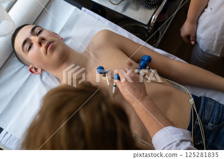 A female doctor performs an ECG on a young man in a hospital room, carefully monitoring his heart health during a routine checkup. A female doctor performs an ECG on a young man in a hospital room, carefully monitoring his heart health during a routine checkup. 125281835