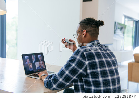 African American businessman in glasses sitting at a table in a modern living room, using a laptop and smartphone for business video chat, conversation with friends and entertainment. African American businessman in glasses sitting at a table in a modern living room, using a laptop and smartphone for business video chat, conversation with friends and entertainment. 125281877