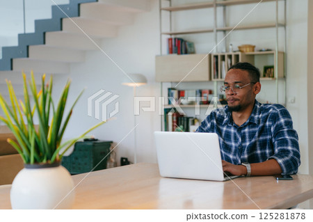African American influencer in glasses sitting at a table in a modern living room, using a laptop for business video chat, conversation with friends and entertainment. 125281878