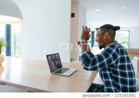 African American businessman in glasses sitting at a table in a modern living room, using a laptop and smartphone for business video chat, conversation with friends and entertainment. African American businessman in glasses sitting at a table in a modern living room, using a laptop and smartphone for business video chat, conversation with friends and entertainment. 125281882