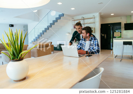 A young married couple is talking to parents, family and friends on a video call via a laptop while sitting in the living room of their modern house. 125281884