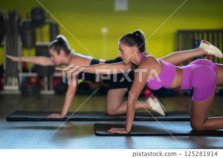 Two fitness women stretch together in the gym, supporting each other in their journey towards flexibility and wellness. 125281914