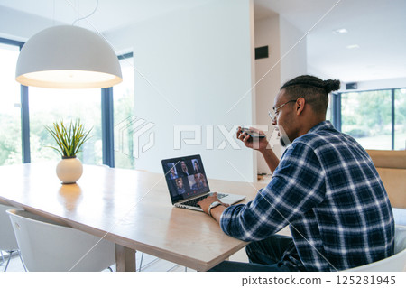 African American businessman in glasses sitting at a table in a modern living room, using a laptop and smartphone for business video chat, conversation with friends and entertainment. 125281945