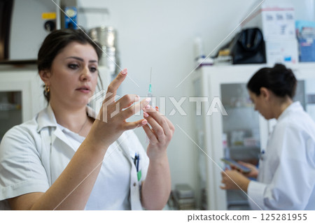 A focused nurse in a white lab coat carefully prepares a syringe for an injection in a professional healthcare environment. 125281955