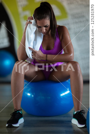 A beautiful and fit woman sitting on a gym ball with a towel, resting and recovering after an intense workout. 125281970
