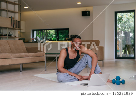 Training At Home. Sporty African American man doing the training while watching an online tutorial on a laptop, exercising in the living room, free space Training At Home. Sporty African American man doing the training while watching an online tutorial on a laptop, exercising in the living room, free space 125281989