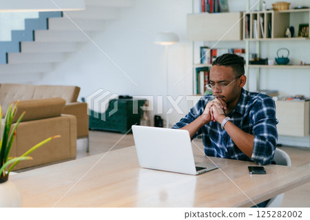 African American influencer in glasses sitting at a table in a modern living room, using a laptop for business video chat, conversation with friends and entertainment. 125282002