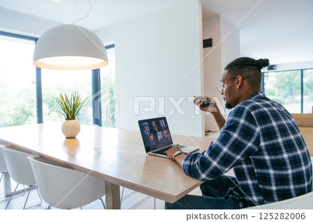 African American businessman in glasses sitting at a table in a modern living room, using a laptop and smartphone for business video chat, conversation with friends and entertainment. African American businessman in glasses sitting at a table in a modern living room, using a laptop and smartphone for business video chat, conversation with friends and entertainment. 125282006