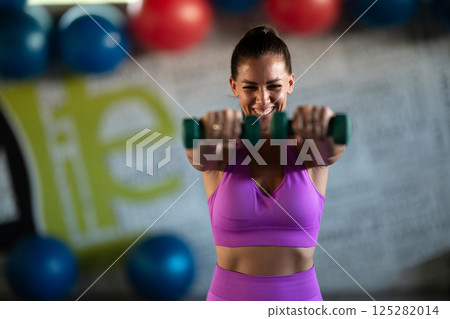 A focused and determined woman performs a strength building dumbbell workout at the gym, showcasing her athletic physique and dedication to fitness. 125282014