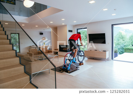 A triathlete riding a triathlon bike on a machine simulation in a modern living room A triathlete riding a triathlon bike on a machine simulation in a modern living room 125282031