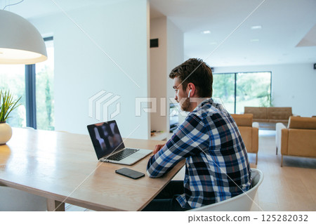 A freelancer sitting at a table in a modern living room, with headphones using a laptop for business video chat, conversation with friends and entertainment A freelancer sitting at a table in a modern living room, with headphones using a laptop for business video chat, conversation with friends and entertainment 125282032