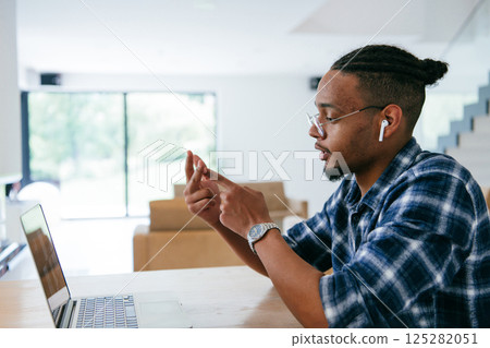 African American influencer in glasses sitting at a table in a modern living room, using a laptop for business video chat, conversation with friends and entertainment. African American influencer in glasses sitting at a table in a modern living room, using a laptop for business video chat, conversation with friends and entertainment. 125282051