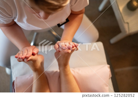 Foot Massage Therapy in a Relaxing Spa Setting. A professional massage therapist in a white uniform provides a soothing foot massage to a client in a calm and serene spa environment Foot Massage Therapy in a Relaxing Spa Setting. A professional massage therapist in a white uniform provides a soothing foot massage to a client in a calm and serene spa environment 125282052