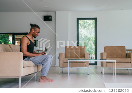 African American man in glasses sitting at a table in a modern living room, using a laptop for business video chat, conversation with friends and entertainment. African American man in glasses sitting at a table in a modern living room, using a laptop for business video chat, conversation with friends and entertainment. 125282053