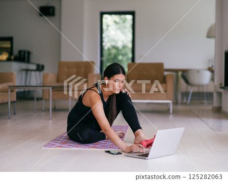 A woman in sportswear is sitting in the living room and preparing for online training while using a laptop. A woman in sportswear is sitting in the living room and preparing for online training while using a laptop. 125282063