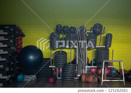 A neatly organized gym equipment storage area featuring a variety of weights, barbells, kettlebells, and fitness accessories against a yellow brick wall. A neatly organized gym equipment storage area featuring a variety of weights, barbells, kettlebells, and fitness accessories against a yellow brick wall. 125282081