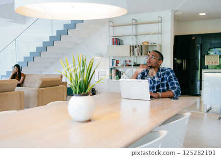 African American influencer in glasses sitting at a table in a modern living room, using a laptop for business video chat, conversation with friends and entertainment. 125282101