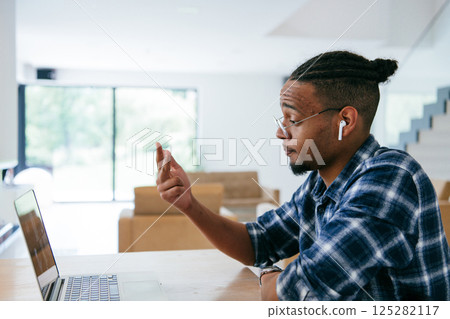 African American influencer in glasses sitting at a table in a modern living room, using a laptop for business video chat, conversation with friends and entertainment. 125282117