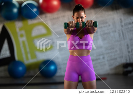 A focused and determined woman performs a strength building dumbbell workout at the gym, showcasing her athletic physique and dedication to fitness. 125282240