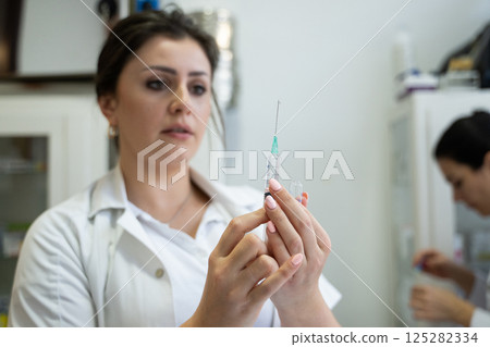 A focused nurse in a white lab coat carefully prepares a syringe for an injection in a professional healthcare environment. 125282334