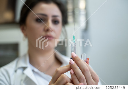 A focused nurse in a white lab coat carefully prepares a syringe for an injection in a professional healthcare environment. 125282449