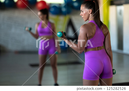 A focused and determined woman performs a strength building dumbbell workout at the gym, showcasing her athletic physique and dedication to fitness. 125282526