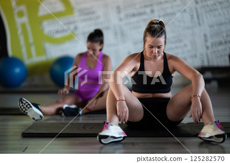 Two fitness women stretch together in the gym, supporting each other in their journey towards flexibility and wellness. 125282570