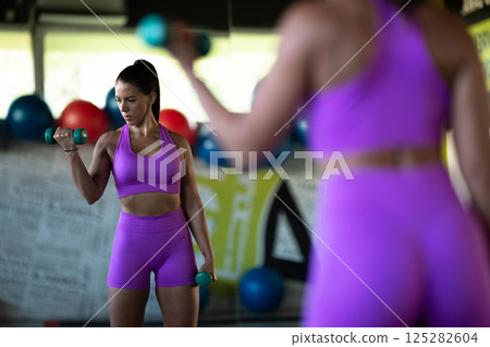 A focused and determined woman performs a strength building dumbbell workout at the gym, showcasing her athletic physique and dedication to fitness. 125282604