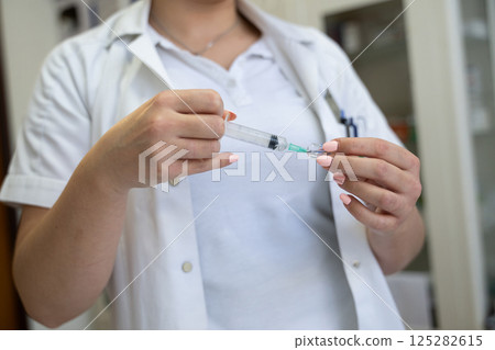 A focused nurse in a white lab coat carefully prepares a syringe for an injection in a professional healthcare environment. 125282615