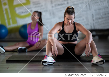 Two fitness women stretch together in the gym, supporting each other in their journey towards flexibility and wellness. Two fitness women stretch together in the gym, supporting each other in their journey towards flexibility and wellness. 125282617